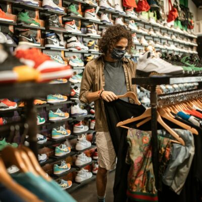 Young man in a sneaker and clothing store wearing a mask, browsing through shirts amid pandemic.